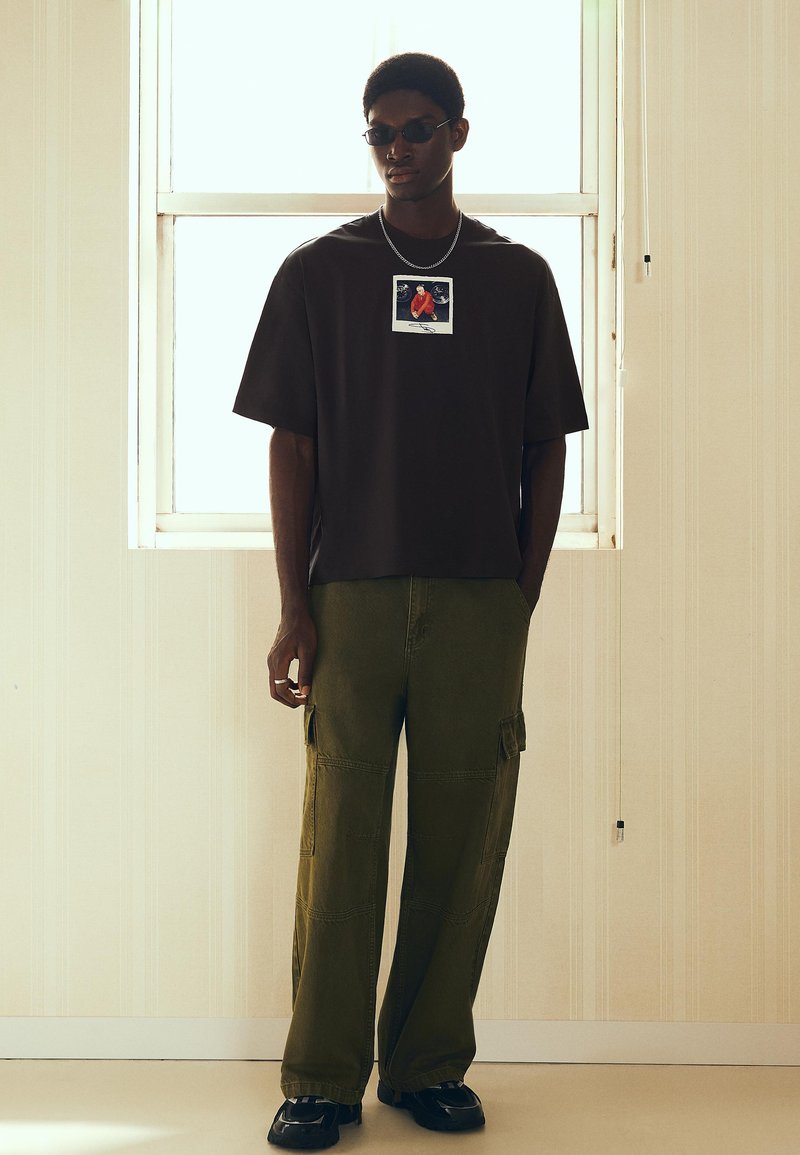 Man draagt een zwart oversized t-shirt met afbeelding, olijfgroene cargobroek, zwarte schoenen en zonnebril, staand voor een raam met tegenlicht in een kamer.