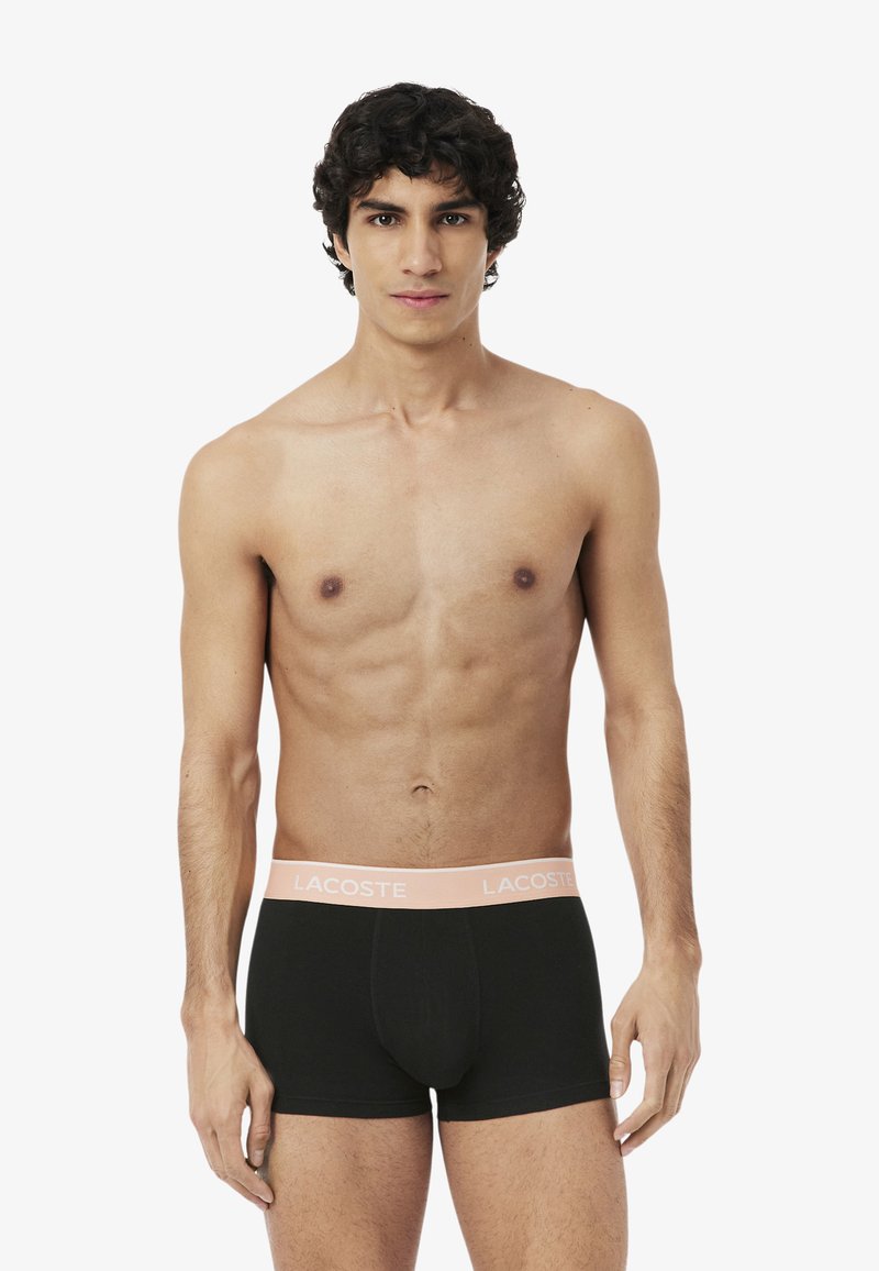 Jonge man met krullend haar, gekleed in zwarte Lacoste boxer briefs met een lichtroze tailleband, staand tegen een witte achtergrond.