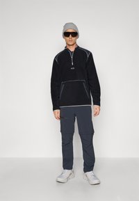 Black fleece pullover with half-zip and grey accents, paired with dark cargo pants featuring multiple pockets. Worn with grey beanie and sunglasses.