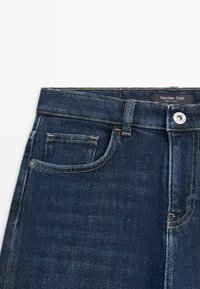 Ciemnoniebieskie dżinsy o średnim stanie, z małą przednią kieszenią, pomarańczowym szwem oraz zapięciem na guzik z srebrnym akcentem.