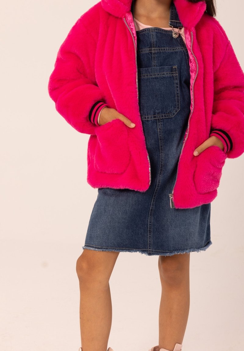 Giacca in pelliccia finta di un brillante colore rosa con polsini a righe bianche e nere, abbinata a un vestito salopette in denim blu con tasca frontale e orlo sfilacciato.
