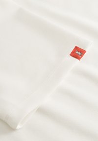 Witte stof met een rood rechthoekig label waarop een kleine witte hondillustratie is geborduurd nabij de zoom.