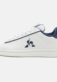 Baskets blanches en cuir lisse, avec un accent en daim marine au niveau du talon. Comprend des lacets assortis et un logo sur le côté.