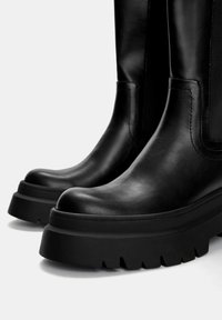 Schwarze Lederstiefel bis zum Knie mit einer dicken, strukturierten Sohle und einer runden Zehenform. Mit glatter Oberfläche und sichtbaren Nähten an den Seiten.