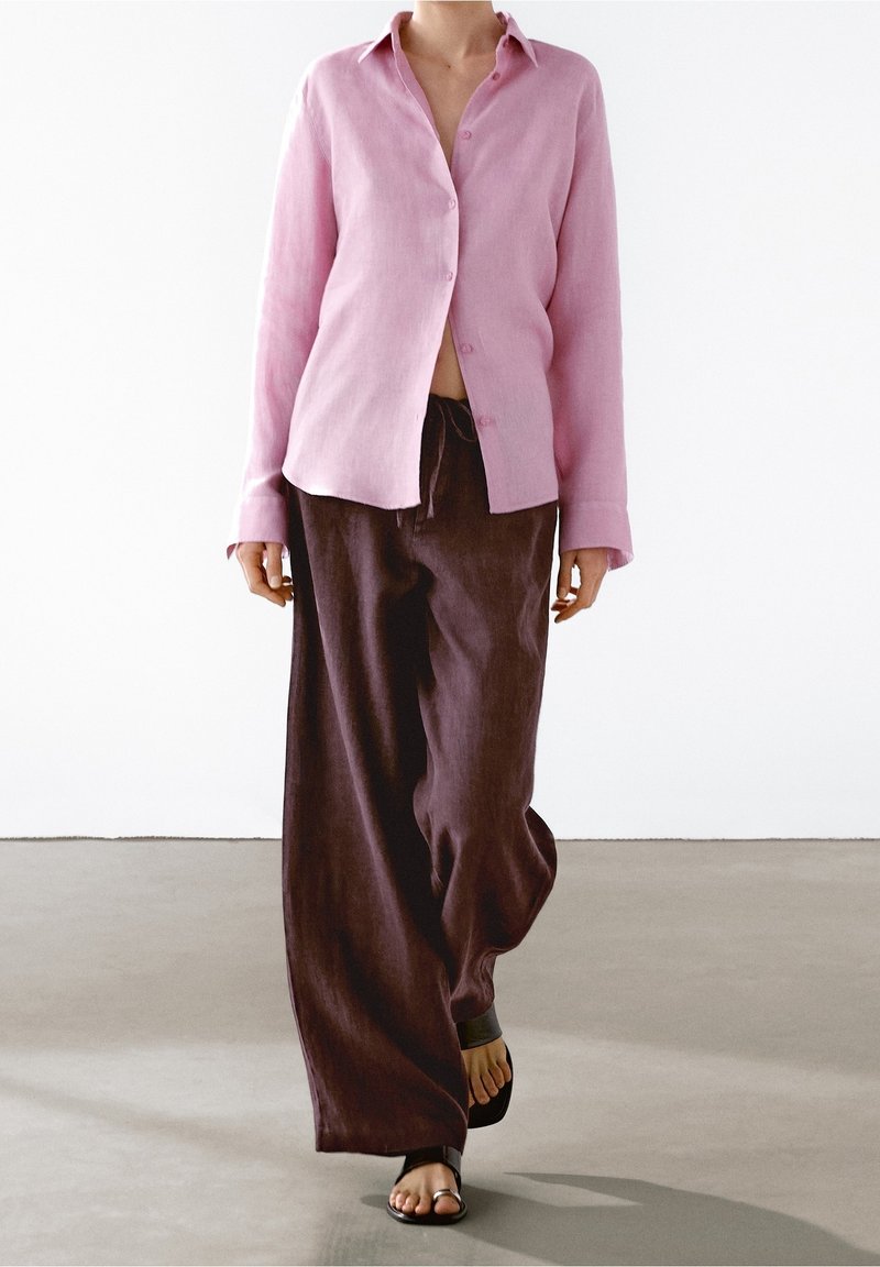 Personne portant une chemise rose clair à boutons, un pantalon large ample marron foncé et des sandales noires à bout ouvert marchant sur un sol gris.