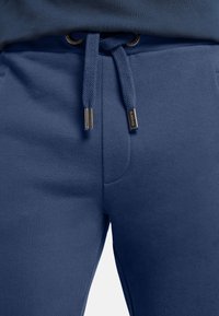 Marineblaue Jogginghosen aus glattem Material mit einem Kordelzugbund mit Metallenden und Seitentaschen. Einfaches, klar strukturiertes Design.