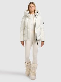 Veste matelassée blanche avec capuche, présentant un motif matelassé en chevrons et une fermeture éclair noire. Associée à des leggings crème et des bottes de neige blanches.