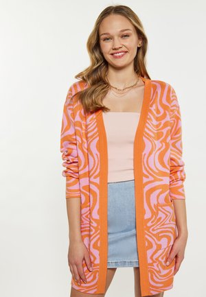 Cardigan lavorato a maglia arancione con motivo ondulato rosa, apertura frontale, maniche lunghe, dotato di tasca, indossato sopra una maglietta rosa chiaro e una gonna di jeans.