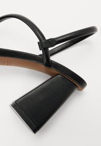 Sandale à talons en cuir noir avec un large talon en bloc texturé. Présente des fines lanières croisées et une semelle intérieure lisse beige.