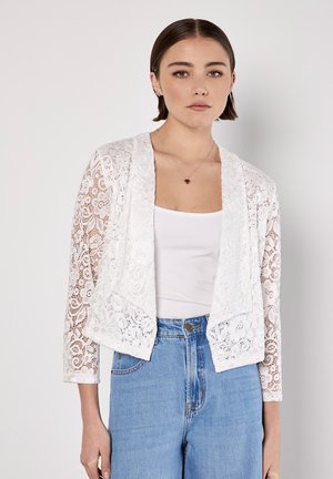 Biały koronkowy kardigan z rękawami o długości trzy czwarte, otwartym kroju, falbaniastymi krawędziami i skomplikowanymi wzorami kwiatowymi, noszony na prostym topie.