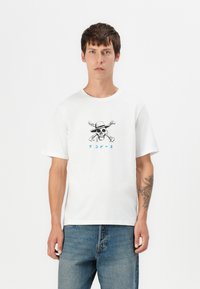 Hvid bomulds-t-shirt med et sort kranie grafik med hat og korslagte knogler, accentueret med blå japanske tekst. Korte ærmer og afslappet pasform.