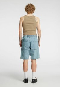 Beige singlet, lyseblå denimshorts med to baklommer, opprullede kanter; hvite sokker og svarte sko synlige. Kort krøllete hår.