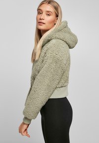 Vezelige, olijfgroene cropped hoodie met een capuchon en elastische manchetten, gecombineerd met zwarte leggings. De stof heeft een zachte, gestructureerde afwerking.