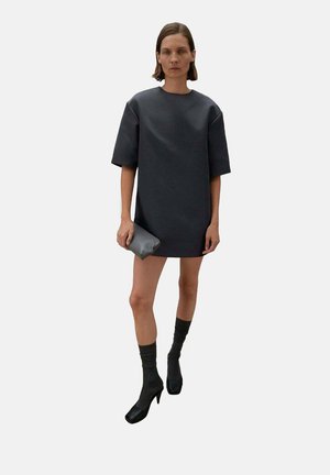 Vestido oversized gris oscuro con mangas cortas, de textura suave. Combinado con zapatos de tacón negro y medias hasta la rodilla oscuras, sosteniendo un pequeño clutch.