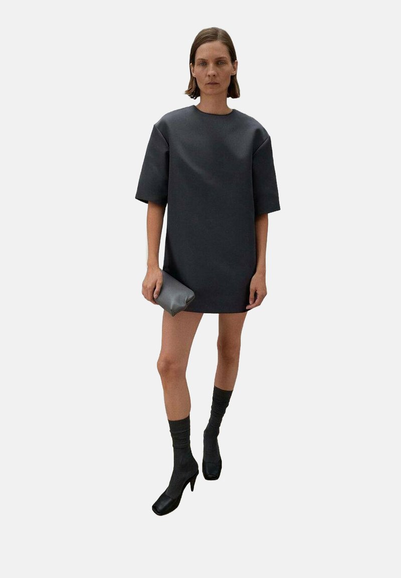Tumma harmaa ylipitkä mekko, jossa on lyhyet hihat, sileä rakenne. Yhdistetty mustiin korkokenkiin ja tummiin polviin saakka ulottuviin sukkiin, kädessä pieni clutch-laukku.