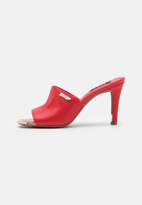Mules ouvertes rouges à talons fabriquées en simili cuir lisse. Présentent un accent logo doré et un talon aiguille avec un détail métallique à la base.