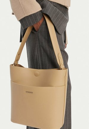 Beige Leder-Schultertasche mit goldenem Logo, gehalten von einer Person, die graue Nadelstreifenhosen und einen beigen Mantelärmel trägt.