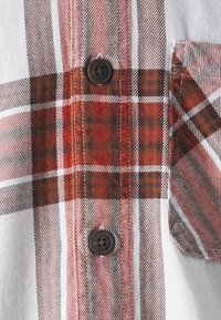 Kostkovaná košile s červenými, hnědými a bílými kostkami. Hnědé knoflíky a náprsní kapsa přidávají designu praktické detaily.