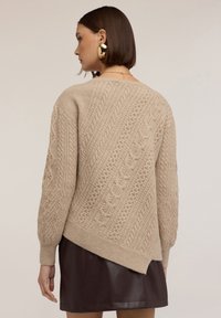 Beige, texturerad tröja med kabelstickad design och ribbade ärmar, med asymmetrisk nederkant, bärs över en mörk läderkjol.