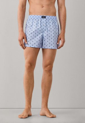 Hombre que lleva puestos unos bóxers azul claro con un pequeño estampado oscuro de remos cruzados, de pie descalzo frente a un fondo gris liso.