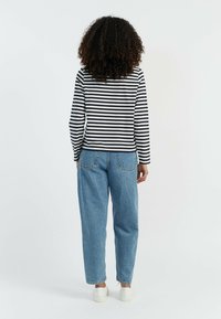 Schwarz-weißtickte langärmlige Bluse, hellblaue hochtaillierte Jeans und weiße Sneaker, von hinten vor einem einfarbigen Hintergrund betrachtet.
