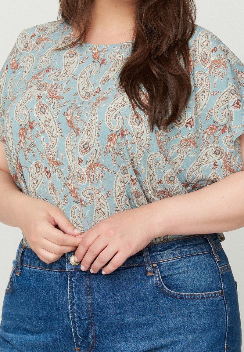 Personne boutonnant une chemise bleue à motif paisley rentrée dans un jean bleu taille haute avec des passants de ceinture visibles.