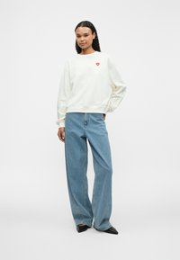 Cream sweatshirt with a red heart logo on the chest, paired with light blue wide-leg jeans and black pointed flats.