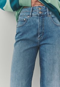 Jean en denim bleu clair avec une taille haute, comportant deux boutons en métal, des coutures contrastées discrètes et un design classique à quatre poches.