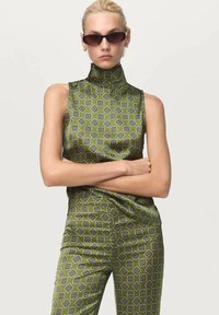 Haut haut en satin verde avec col montant et pantalon assorti à un motif géométrique. Présente une silhouette ajustée et un design épuré. Lunettes de soleil noires portées.