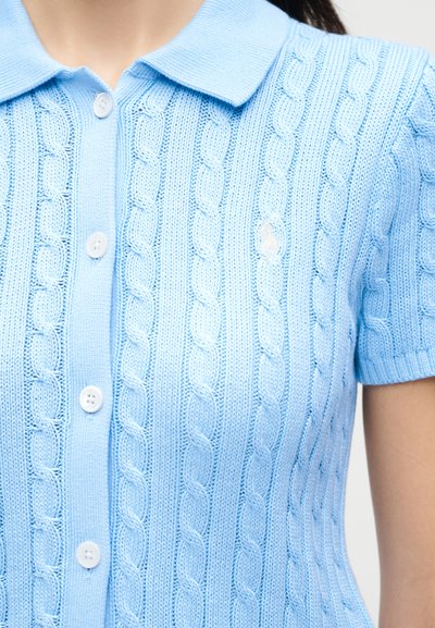 Gaiši zils īsroku polo krekls ar adījuma rakstu, baltām pogām un apkakli, valkāts uz personas ar redzamu kaklu un roku.