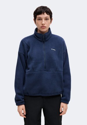 Jeune personne aux cheveux courts et foncés portant une veste polaire bleu marine avec une fermeture éclair quart et un pantalon noir, debout face à l'avant.