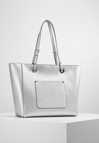 Srebrna torba na ramię z teksturowanym wykończeniem, z dwoma ramiączkami, przednią kieszenią i metalicznymi akcentami. Prostokątny kształt.