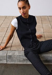 Schwarze gestrickte ärmellose Weste mit Knöpfen, getragen über einem weißen kurzärmeligen Hemd. Das Model sitzt auf einer gefliesten Fläche und trägt gestreifte schwarze Hosen.