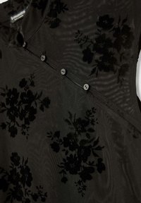 Schwarze Bluse mit floralen Mustern und transparenter Textur, die ein dezentes, tonales Muster aufweist, sechs kleine Knöpfe hat und ein schräges Design an einem Schulterteil hat.