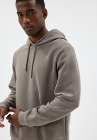 Grijze hoodie van zachte stof, met een trekkoordkap, geribbelde manchetten en een relaxte fit, gedragen door een model tegen een neutrale achtergrond.