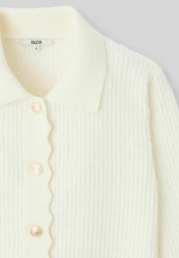 Cardigan crème côtelé avec une patte de boutonnage festonnée et des boutons dorés, doté d'un col et d'une texture douce. Taille S.