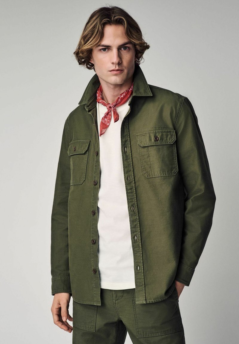 Olive green shirt jacket with two chest pockets and brown buttons, worn over a white shirt, complemented by a red bandana.