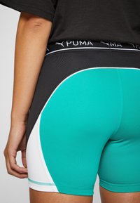 Zbliżenie na osobę w turkusowych, czarnych i białych szortach sportowych Puma oraz czarnej koszulce, ręka luźno opuszczona przy boku, widok z tyłu.