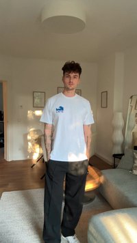 T-shirt branca de algodão com gráfico de onda azul, combinado com calças pretas. De pé numa sala bem iluminada com piso de madeira clara.