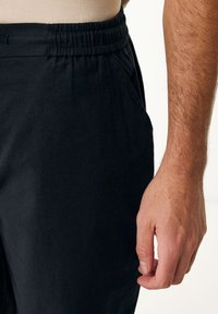Pantalones negros de cintura elástica con bolsillos laterales, fabricados con un material ligero y un corte relajado. Mano reposando en el costado con detalles de textura visibles.