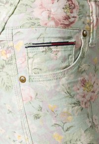 La tela de denim verde con estampado floral presenta flores en tonos rosa, amarillo y blanco. Incluye un pequeño bolsillo con acentos a rayas rojas y marinas.