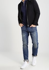 Svart zip-up jacka över en marinblå polo, kombinerad med ljusblå slitna jeans och vita sneakers, med uppvikta ärmar och en fickdetalj.