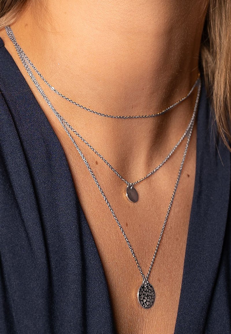 Tre collane d'argento a strati: delicate catene con pendenti ovali e texturizzati. Le collane variano in lunghezza e brillano contro una maglietta scura.