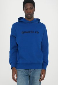 Felpa blu realizzata in tessuto morbido con tasca a marsupio frontale. Presenta la scritta "QUARTZ CO" in nero sul petto. Design semplice a pullover.
