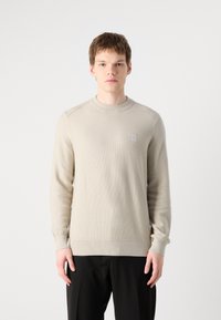 Beige neulottu sweatshirt, jossa on ribbardettu pinta, pyöreä pääntie ja pieni logo-patch, yhdistetty mustiin housuihin.