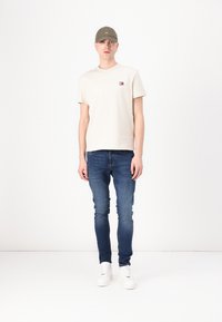 Beige bavlněné tričko s malým barevným logem, nošené s tmavě modrými slim-fit džíny a bílými teniskami. Olivově zelená kšiltovka doplňuje vzhled.