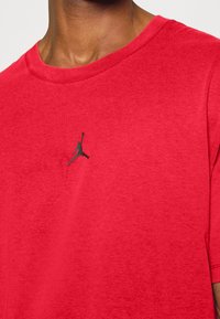 Camiseta roja de algodón suave con cuello redondo, mangas cortas y un logo Jumpman negro en el pecho. Textura lisa, diseño casual.