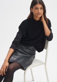 Maglione oversized nero a maglia con collo alto, abbinato a una gonna nera in pelle trapuntata. Modello seduto su una sedia bianca, con indosso collant neri.