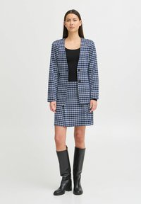 Blazer à carreaux bleu et blanc avec jupe assortie, comportant deux poches avant, associé à des bottes noires jusqu'aux genoux et un haut noir ajusté.
