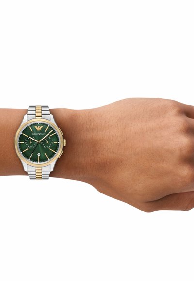 Handgelenk mit zweifarbiger Metallarmbanduhr mit grünem Zifferblatt, goldenen Akzenten, drei Unterzifferblättern und einem Datumsfenster bei der Sechs-Uhr-Position.